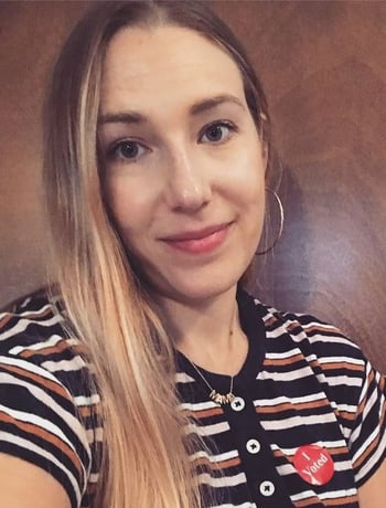 Adrienne Aarts smiling against a wooden background, wearing a black, orange, and white button-up shirt, an "I Voted" sticker, a small gold necklace, and hoop earrings.