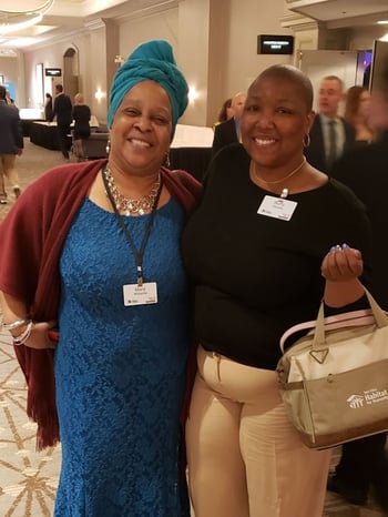 Buffy Moore smiling with her mother in a hotel hallway. Buffy in a black shirt and tan pants with a beige Habitat bag and nametag, plus a gold necklace and earrings. Her mother in a blue lace dress, red shawl, blue headwrap, nametag on a lanyard, and silver necklace, earrings and bracelets.