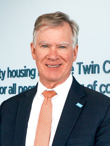 Chris Coleman smiling and standing in front of blue text on a white wall that shows the TC Habitat mission statement. Wearing a dark suit with a light blue Habitat pin, white shirt, and peach tie with a small pattern on it..