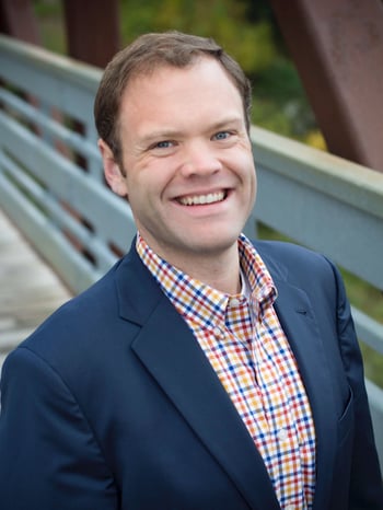 Jim Mulrooney standing on a bridge and smiling, wearing a dark blue suit jacket with a yellow, red and blue plaid shirt.