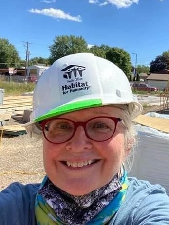 Karen Welle, Volunteer, smiling and standing outside a build site in the sun, wearing glasses, a white hard hat, a light blue sweater jacket, a blue and green neck scarf, and a gray and white patterned mask around her neck.