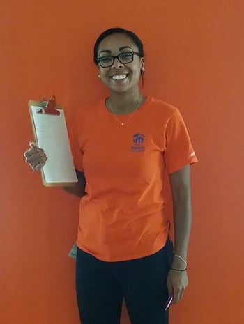 Mari Ojeda smiling and standing in front of an orange background while holding a clipboard in one hand. She's wearing glasses, earrings and a necklace with two small bracelets, an orange TC Habitat shirt, and dark pants. In her other hand she holds a pen, and a nametag is clipped to her left hip.