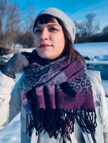 Polina Montes de Oca outside in the snow and looking off into the distance, wearing a light gray hat and vest, darker gray gloves, a white cable knit sweater, and a purple and gray leopard-print scarf.