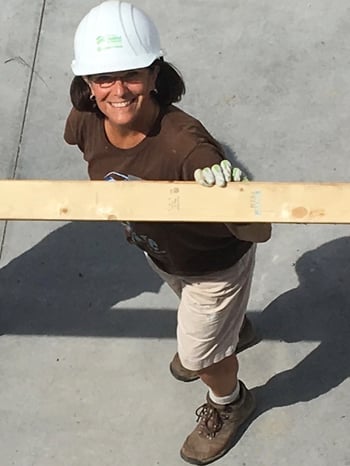 Sharon Mason, standing on concrete below the camera and looking up and smiling. Wearing a brown t-shirt, khaki shorts, work boots, gloves, a white hard hat, and glasses. She's holding a wooden beam.