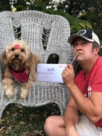 Skip Schmall, Site Supervisor, outdoors and wearing khaki shorts and a red t-shirt with a black and white baseball cap. He is kneeling while looking ponderous holding a pen to his chin, by a gray wicker chair that has his dog, Maple, sitting atop it, with an "I Voted" sticker decorating her forehead and a black and red checkered bandana around her neck. She sits next to an election ballot in its envelope, which is standing up.