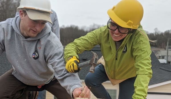 An AmeriCorps member on the right, she's wearing a green jacket with the AmeriCorps logo, a yellow hard hat, safety goggles, and yellow gloves as she uses a hammer on a roof. To her left is another member, he's in a gray AmeriCorps sweatshirt, holding the nail, wearing a white hard hat.