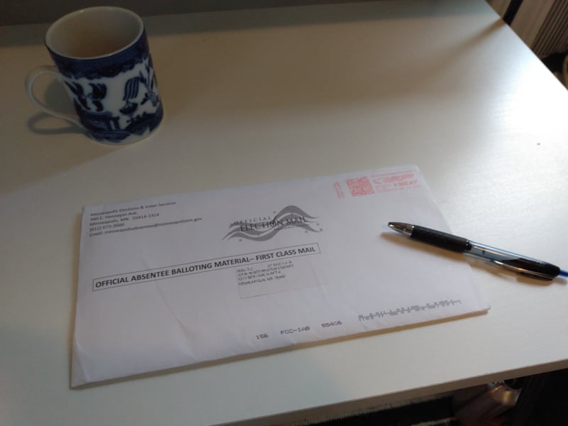 On a white desk, an official absentee ballot envelope, with a black pen lying on top of it. There's a white mug with blue designs on it in the upper left corner of the desk.