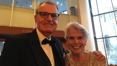 Bill Sands in a black suit and tie, Susan Sands in a beige embellished top, smiling.