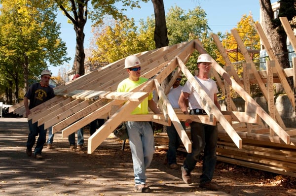 Hard hat works carrying the frame of a roof of a house.