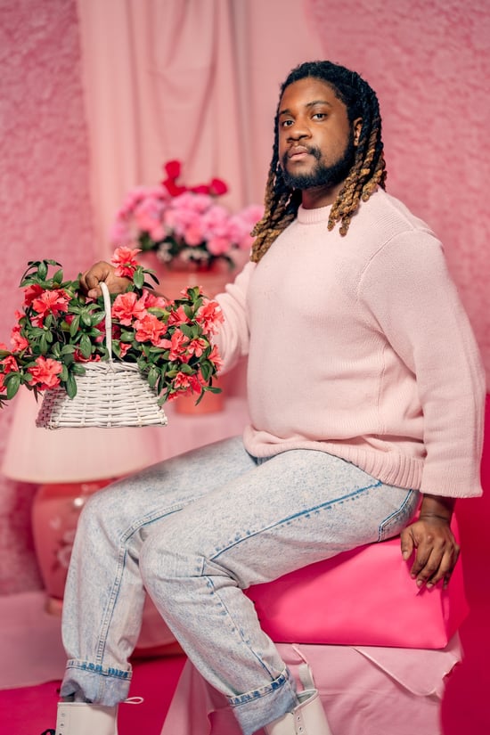 Denzel, a queer Black man, posing in front of a pink background and holding a basket of pink flowers.