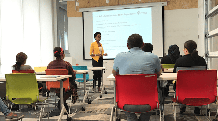 Red, green, orange, and blue chairs in a classroom at six tables, the chairs are mostly filled. At the front of the room is a woman gesturing as she teaches, wearing a white shirt, yellow jacket, black pants, and a bracelet. Behind her is a powerpoint presentation with the title "The Role of a Realtor in the Home Buying Process" and TC Habitat's logo.