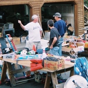 Dennis in a white shirt and khakis in front of his garage, gesturing with one arm. Two men in blue shirts and jeans (one in a baseball cap) stand listening to him. Surrounding them on the driveway are multiple tables covered in boxes and miscellaneous items for donation to ReStore.