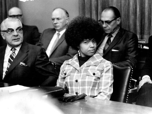 Josie Johnson in a black and white photo, sitting at a table in a conference room for the Board of Regents. She's wearing a plaid overcoat and earrings, a briefcase and gloves sitting on the table in front of her. The other seats in the room are filled by white men in suits.