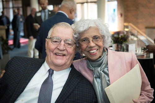 Walter Mondale and Dr. Josie Johnson smiling in the atrium of the University of Minnesota School of Public Affairs. Mondale is wearing a dark plaid suit jacket, glasses, a blue patterned tie and a white shirt. Dr. Johnson is wearing glasses, dangly earrings, a light pink suit jacket, and a silver blouse while holding a folder.