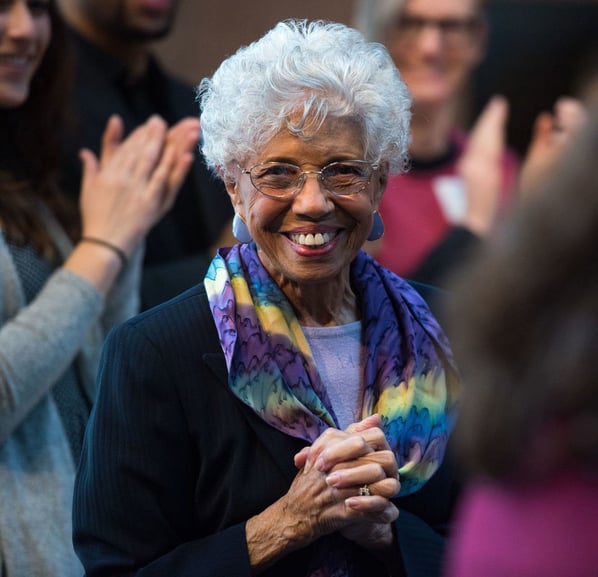 A close-up on Dr. Josie Johnson, in a purple shirt, black pinstripe jacket, and a blue, purple and yellow patterned infinity scarf, smiling in a crowd with her hands clasped.