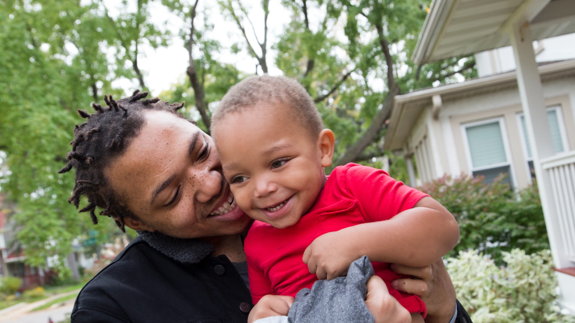 A parent with short hair smiling while holding their child next to their house.