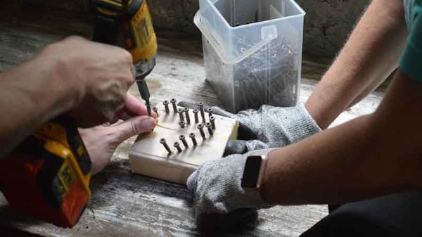 On a wooden floor, nails are being screwed into a block of wood in a heart shape by two people who are out of frame except for their arms. The one on the right has gray work gloves and a smartwatch. A clear plastic box of screws is visible above the block of wood.