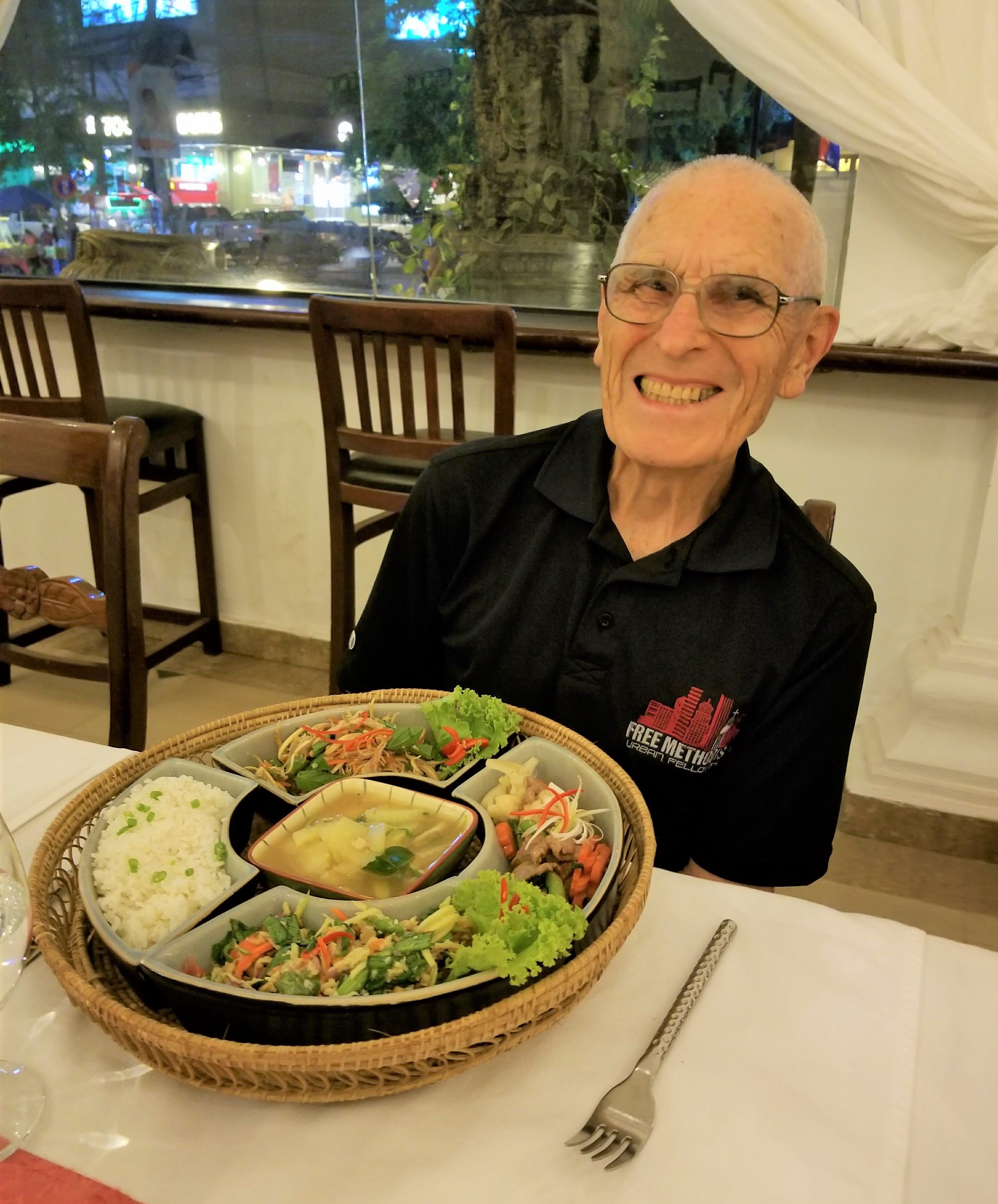 A smiling Orin is seated at a Cambodian restaurant with a plate of varied foods in front of him. 