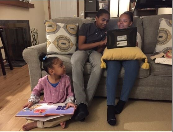 Don helping his sister with homework on the couch while his younger sister looks up at them from the floor, holding a book.