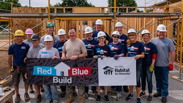 Members from both parties of the Minnesota Legislature, wearing hard hats and gloves, standing in front of the frame of a house in the sunlight and holding a sign that says "Red Blue Build, Twin Cities Habitat for Humanity."