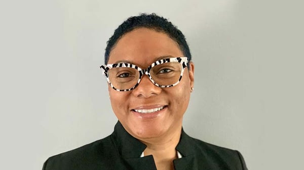 Shereese Turner smiling in front of a gray background, wearing a white shirt, black formal jacket, and black and white striped glasses.