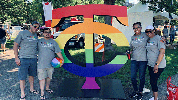 A rainbow sign saying "TC" with the T intersecting the C. Four Habitat employees in gray t-shirts that say "Build Pride" with a rainbow on them stand flanking the sign. They are outside at TC Pride festival.