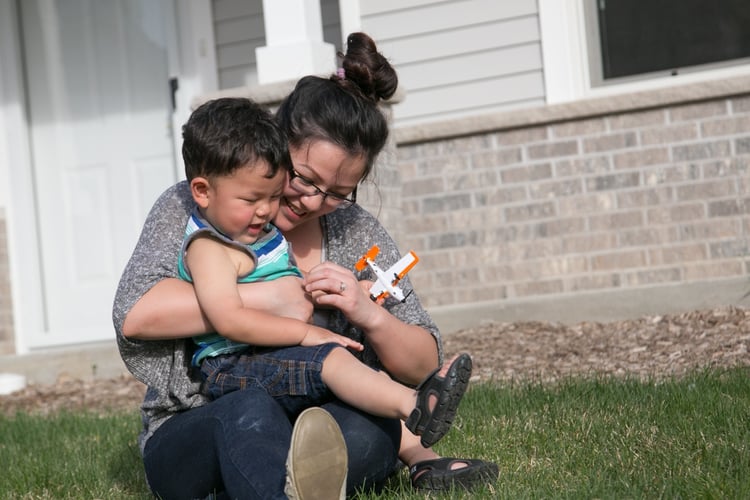 A Habitat homeowner laughing while holding her son in their front yard. She's wearing glasses, a gray top and jeans, and her son is in a blue, green, and white striped shirt and jean shorts with sandals. Her son is holding an orange and white airplane.