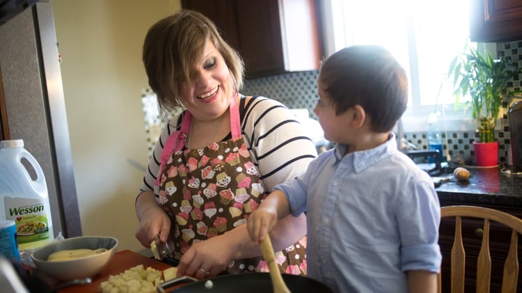 A Habitat homeowner smiling with her son as they cook at the counter of their kitchen. She is wearing a black and white striped shirt with a pink and brown apron covered in cupcake designs, and her son is standing on a chair in a light blue button-up shirt as he stirs a spoon in a pan. The homeowner is cutting vegetables next to a bowl, and a variety of ingredients.