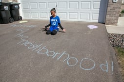 "Welcome to the neighborhood!" written on a driveway, with the artist kneeling next to it.