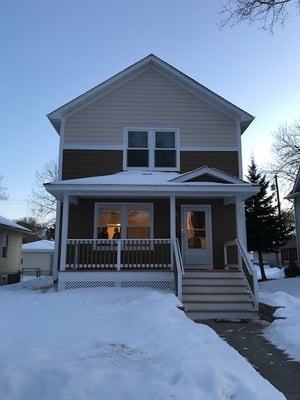 With a clear sky in the snow, a two-story Habitat house with peach siding at the top near the roof, and brown siding on the bottom half of the house. A short set of stairs leads up to the covered porch and front door. The house has a pitched roof and white trim.