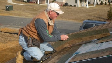 A man in a white hat, gray long-sleeved shirt, brown vest, work books, and jeans kneels on a roof of a house to inspect it.  