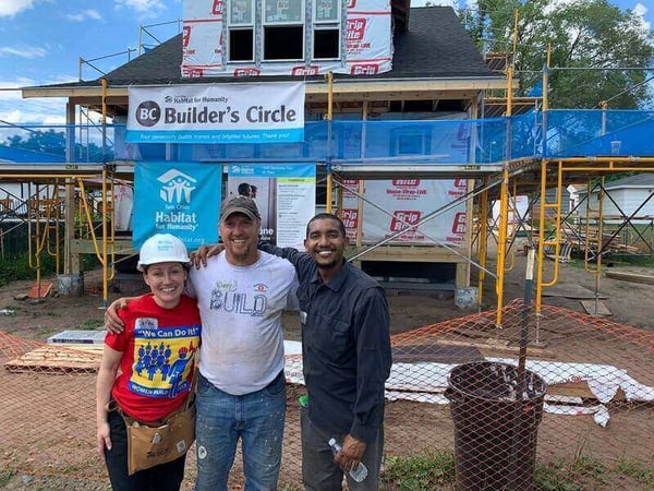 Corey, Catina, and Abdi standing outside a partially-finished home with the Builder's Circle and TC Habitat signs behind them.