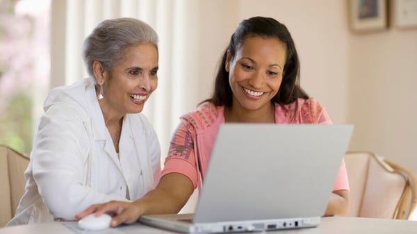 A mother and daughter smiling and sitting at a table looking at a laptop.