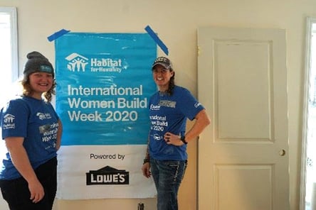 Two women in blue Women Build Week t-shirts next to a blue sign that has the TC Habitat logo and the text "International Women Build Week 2020". Below the text is a section that says "Powered by Lowe's".