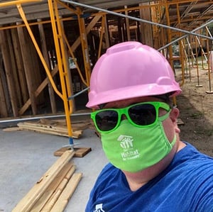 A volunteer in front of a framed house, wearing a green mask and sunglasses, and a pink hard hat.