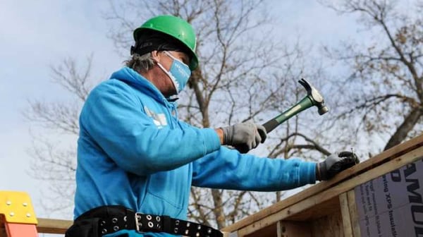 Chris coleman swinging a hammer outside a Habitat house in a blue sweatshirt, green hard hat, and face mask