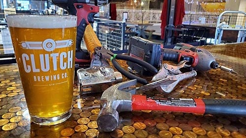 A glass of beer with the Clutch Brewing logo, along with a red hammer, power screwdriver, pliers and a tape measure, all sitting atop a tabletop made of coins in the middle of a bar.