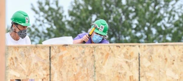 Two volunteers working behind a wall. The one on the right uses a yellow hammer while wearing a purple shirt, green hard hat, and surgical mask. The one on the left watches while wearing a white shirt, green hard hat, and black and white patterned mask.