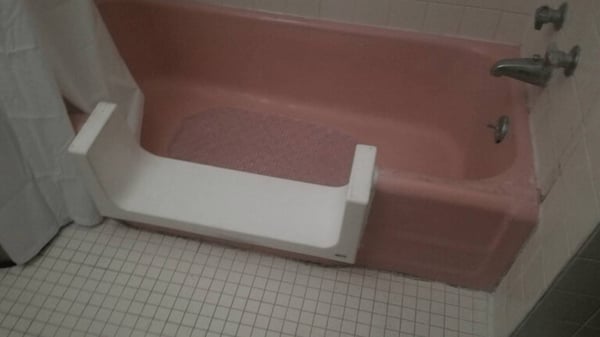 White tile in a bathroom, surrounding a pink bathtub. The middle of the side of the tub has been cut out, and replaced with a u-shaped white piece so you can step into the tub.