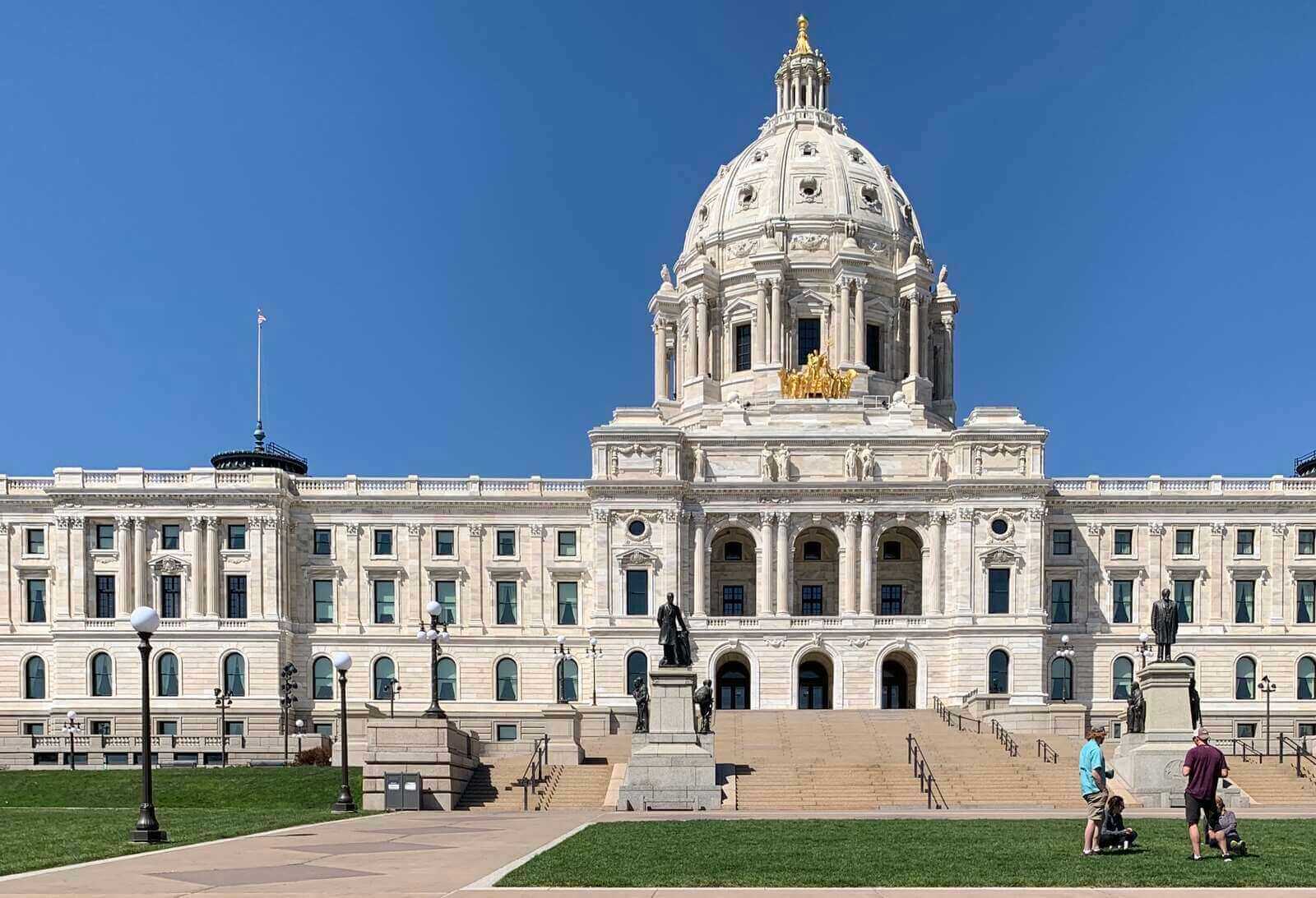 A view of the front of the Capitol Building, on a bright sunny day with green grass, and a couple of people with strollers on the lawn.