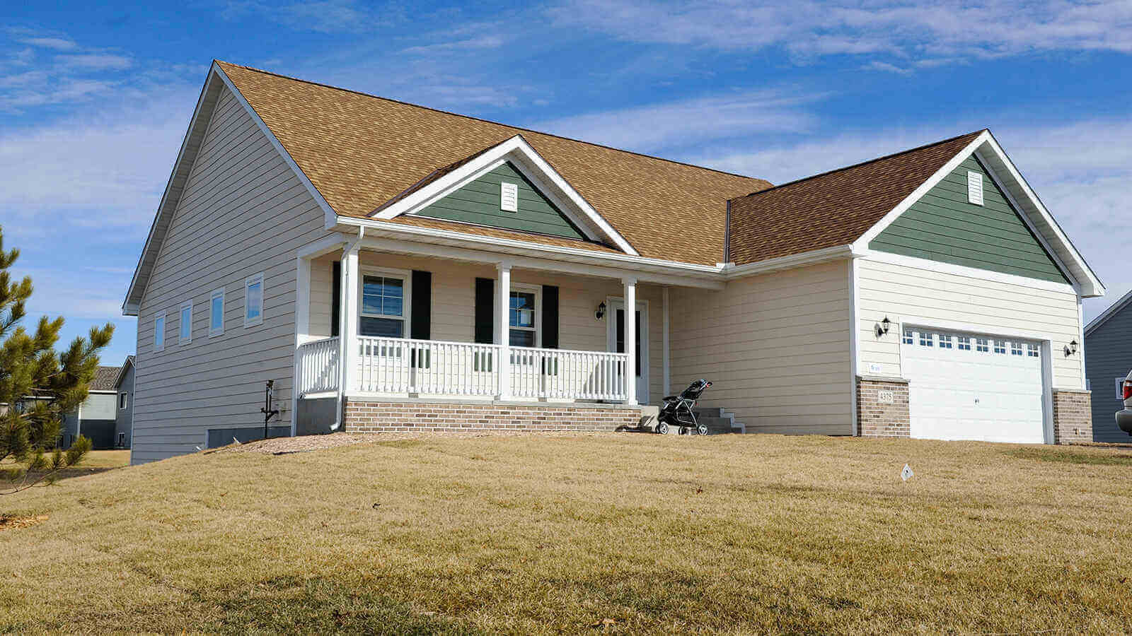 A pale peach home with a garage attached on the side. The areas directly under the roof are painted hunter green, as well as the window shutters. There is white trim and a brown roof, as well as some decorative brick around the base of the garage and front porch.