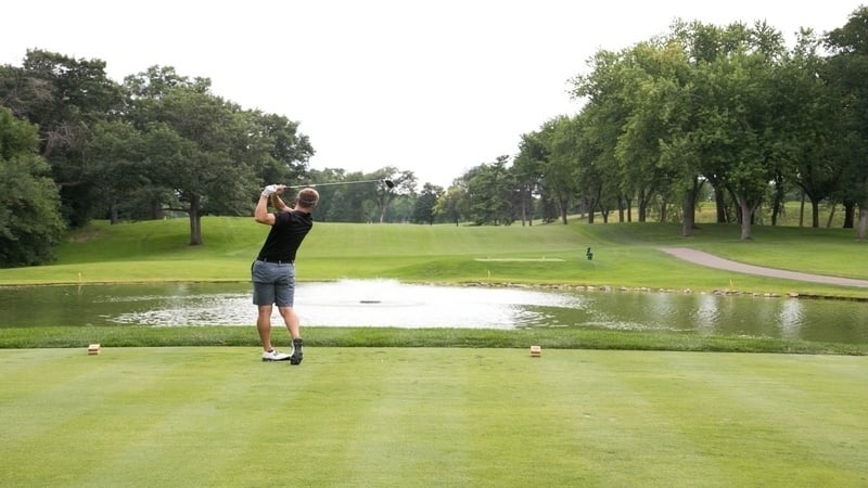 A man in a black shirt and gray shorts swinging a golf club in front of a pond on a golf course, on a cloudy day.