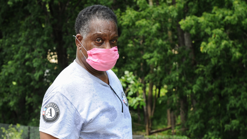 Warren outside in front of a group of trees, facing the camera in a gray AmeriCorps t-shirt, wearing a pink mask.