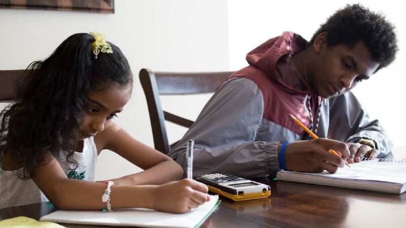 Two siblings sitting at a kitchen table doing homework with a calculator.