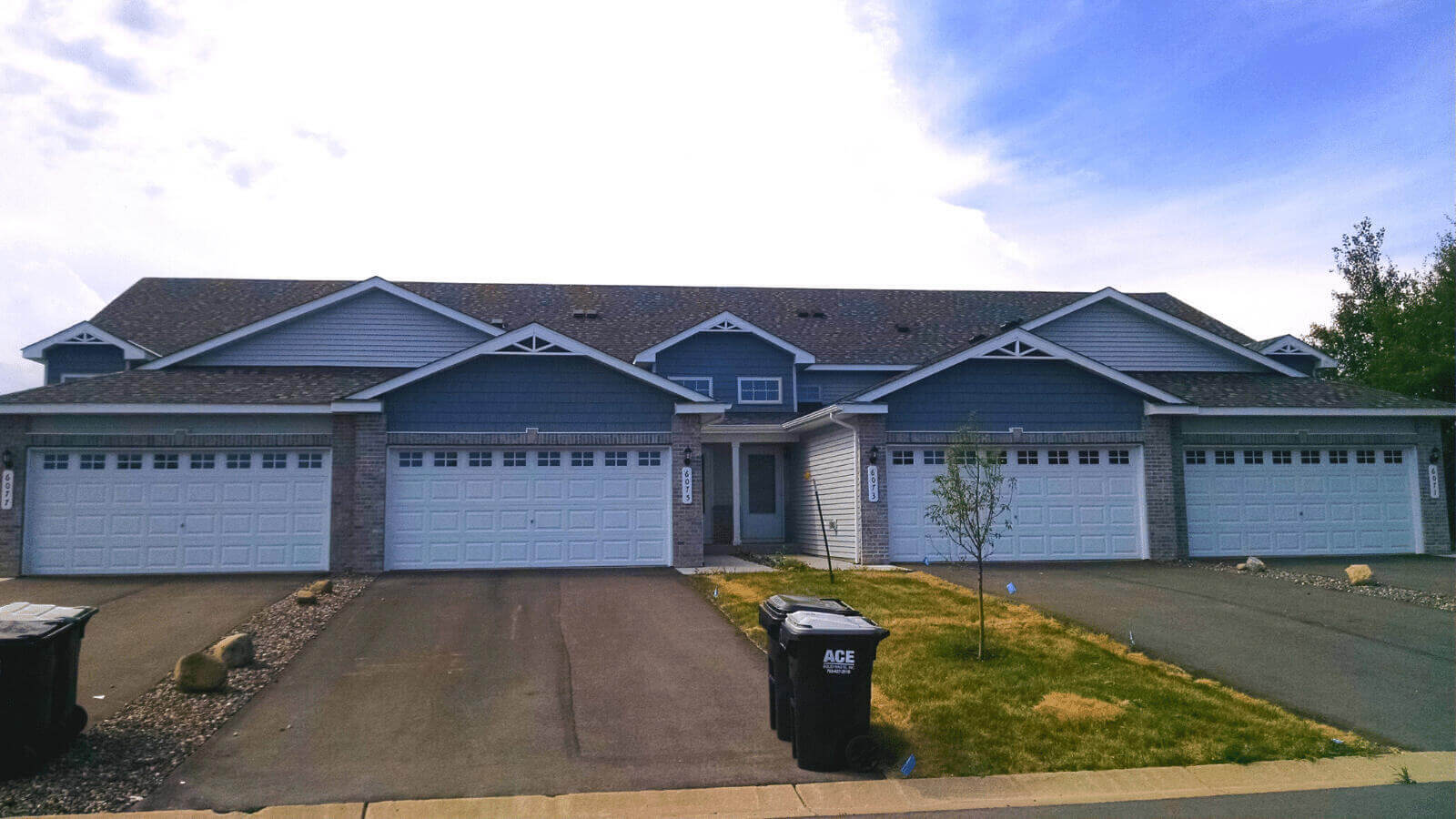 Two gray townhouses side-by-side in the sunlight. A waste bin and recycling bin sit in the front of one of the driveways.