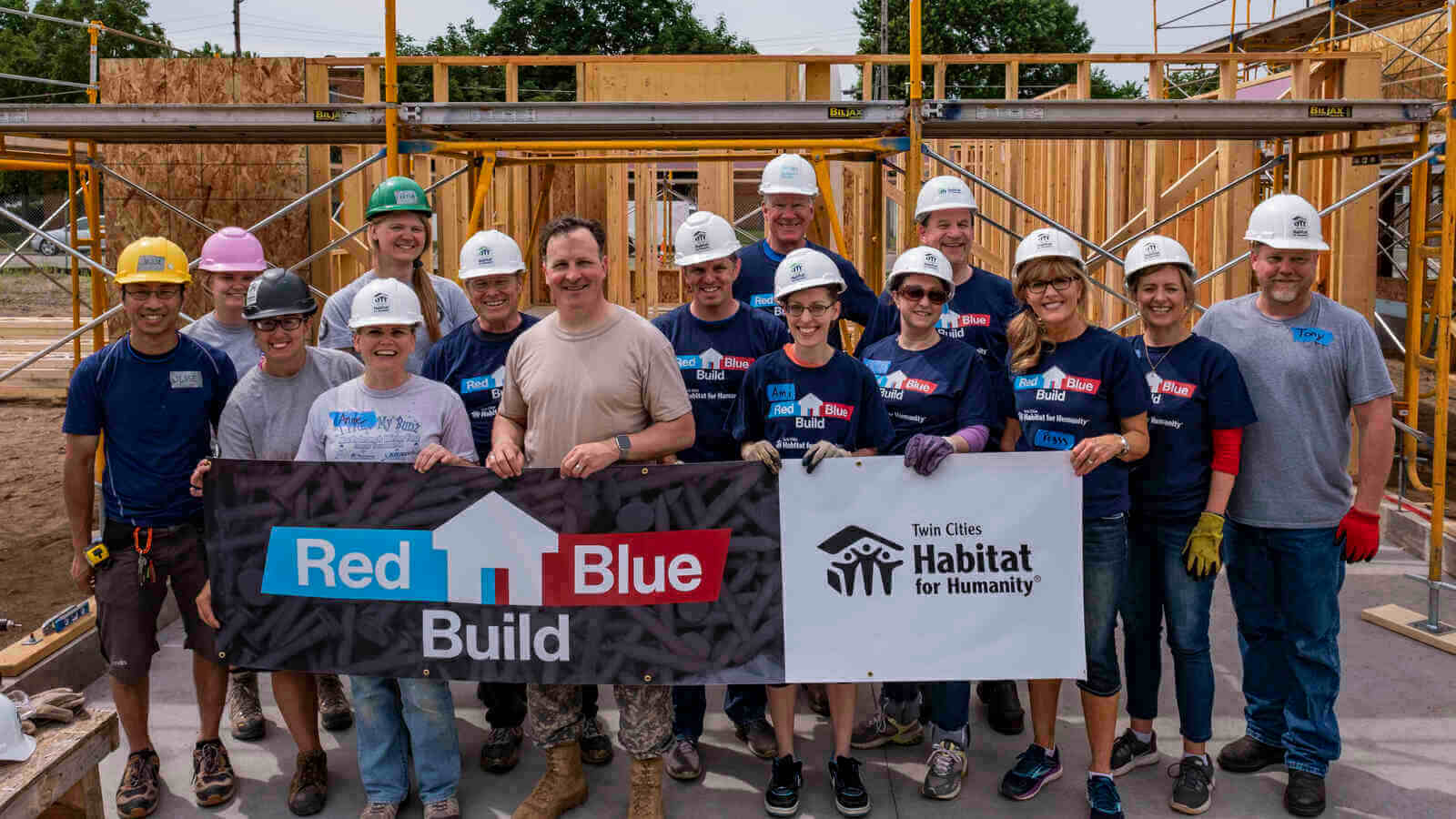 Members from both parties of the Minnesota Legislature, wearing hard hats and gloves, standing in front of the frame of a house in the sunlight and holding a sign that says 