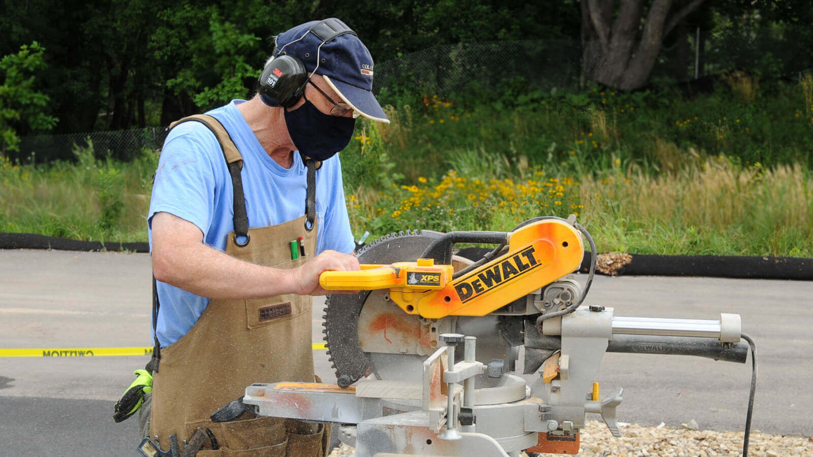 volunteer standing outdoors in a light blue shirt, tan overalls, blue baseball cap and a black mask, wearing ear protection while using a power saw.