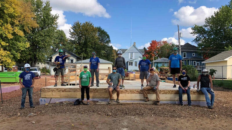 Eleven volunteers standing and sitting on a recently-constructed floor at a build site between several houses.