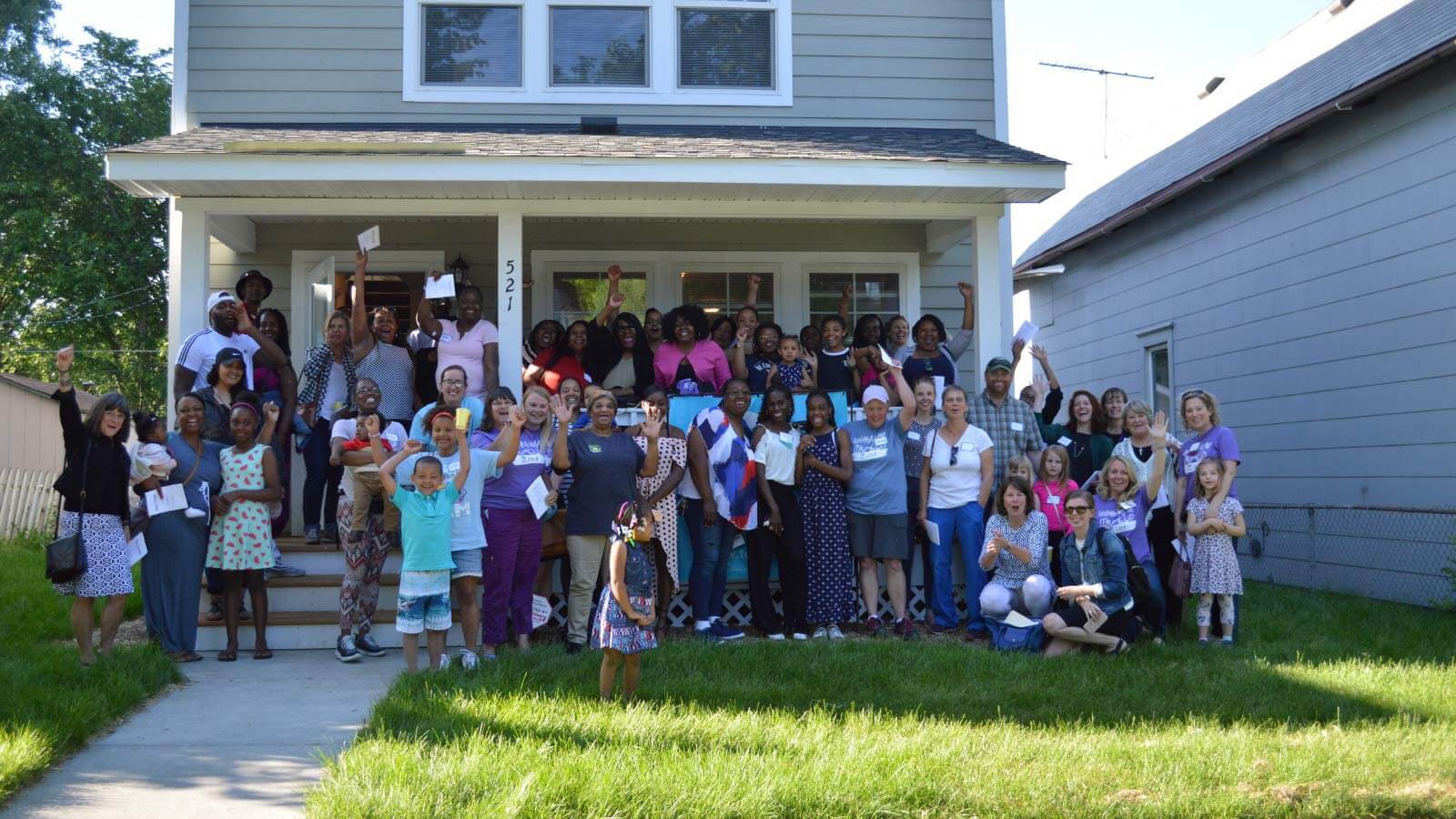 Dozens of people smiling and waving outside of LeAndra's home, with many people on the front porch and many on the lawn in the shade.