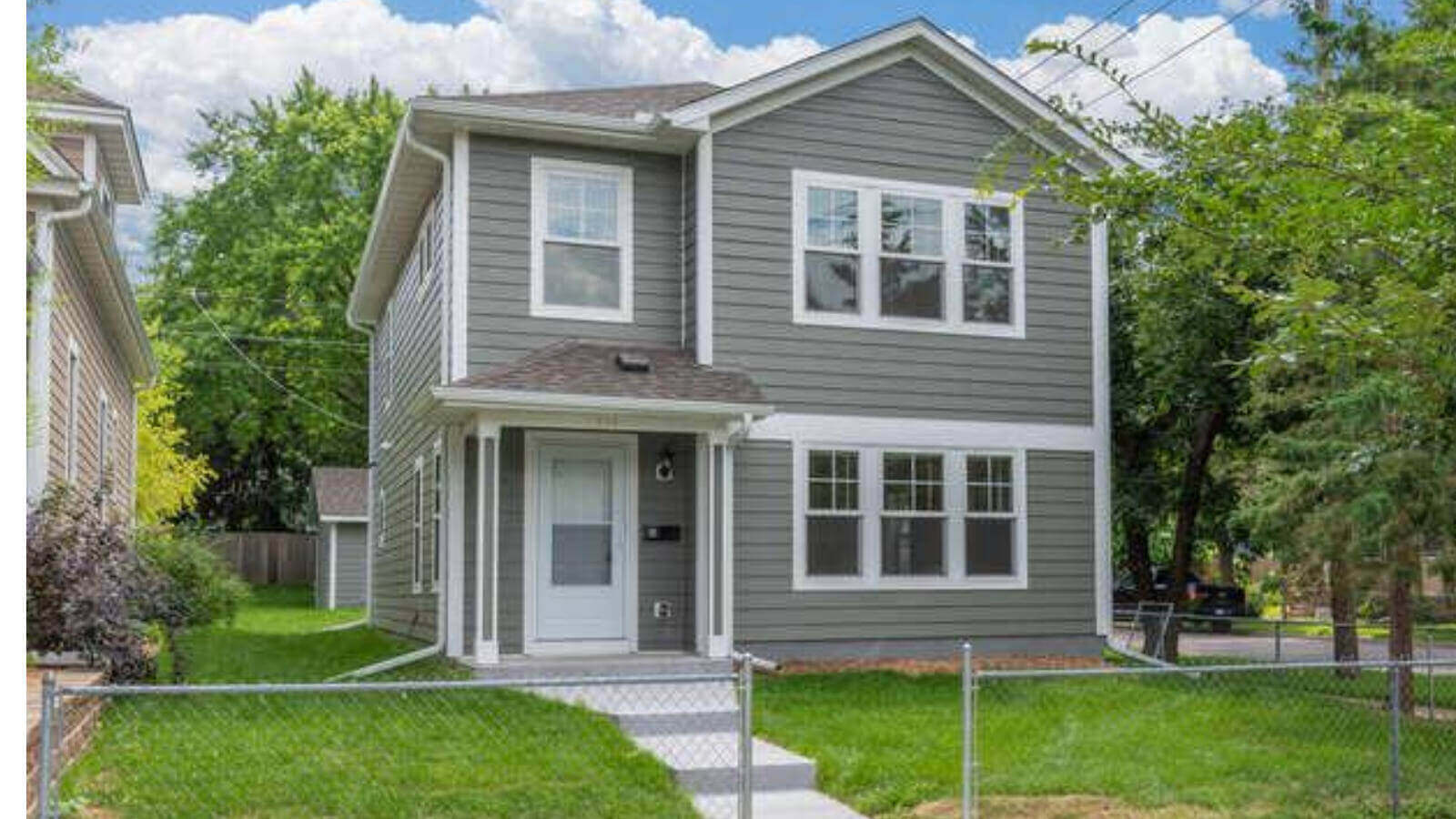 The House that Beer Built, a gray two-story house sitting on a green lawn with trees to the right, a wire fence at the front, and concrete steps leading down to the fence.